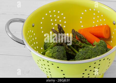 Washed asparagus, broccoli and carrot vegetables in a bright green collander strainer.  With white wood background Stock Photo