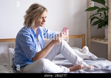 Blonde woman using smartphone sitting on bed. Reading news and online books. Social media, instagram, stay home and quarantine Stock Photo