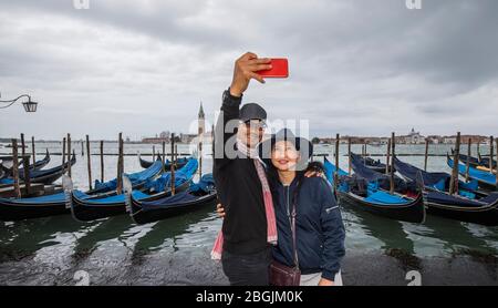 Thai couple taking selfie in front of parked gondola's in Venice Stock Photo