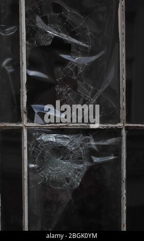 Close up of two window lights with circular impact damage to the glass, and reflections on the uneven surface.Broken panes, shards and frame:Vandalism Stock Photo