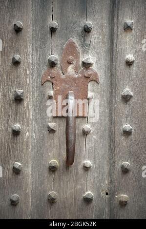 Rusty old fleur-de-lis door handle on cracked wooden door panel studded with rows of wooden beads. Stock Photo