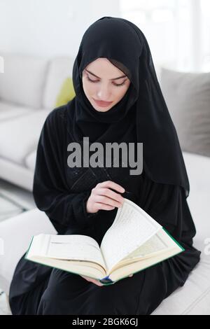young traditional muslim woman reading Quran on the sofa before iftar dinner during a ramadan feast at home Stock Photo