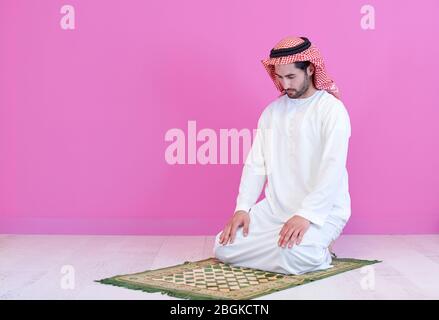 young arabian muslim man in traditional clothes praying on the carpet in front of pink wall before iftar dinner during a ramadan feast at home Stock Photo