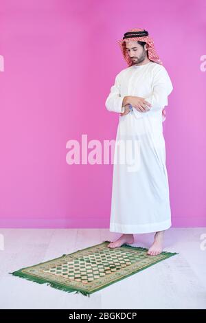 young arabian muslim man in traditional clothes praying on the carpet in front of pink wall before iftar dinner during a ramadan feast at home Stock Photo