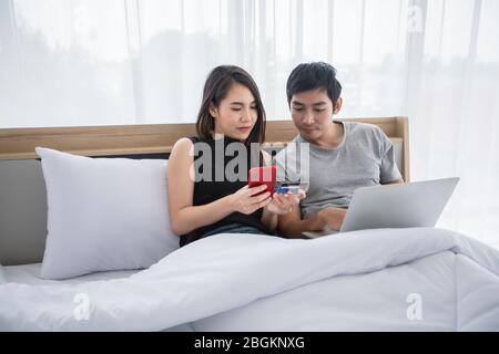 Asian Couple shopping online and paying with credit card on laptop and mobile phone at home,Happy couple at home surfing the net in bed Stock Photo