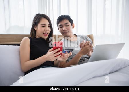 Asian Couple shopping online and paying with credit card on laptop and mobile phone at home,Happy couple at home surfing the net in bed Stock Photo