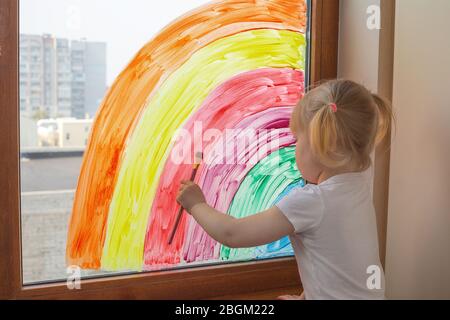 Caucasian little child girl draws a rainbow on the window Stock Photo
