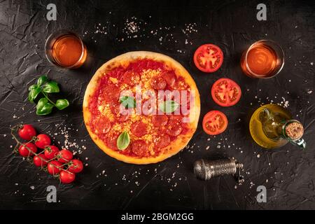 Pepperoni pizza on a black background, with wine glasses and ingredients, tomatoes, basil, and olive oil, shot from the top Stock Photo
