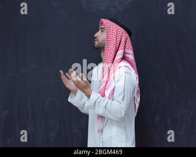 young arabian man in traditional clothes making traditional prayer to God, keeps hands in praying gesture in front of black chalkboard representing mo Stock Photo
