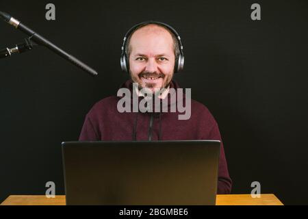 Guy in hoodie sitting behind the desk with laptop computer and headphones on black background, talking to microphone, recording video blog Stock Photo