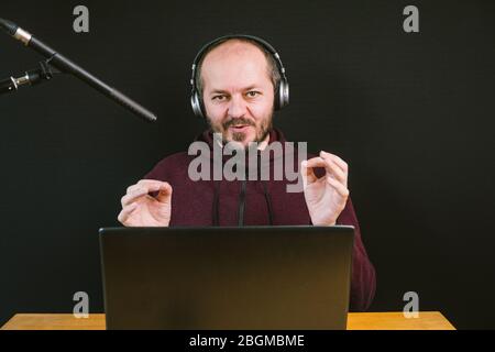 Guy in hoodie sitting behind the desk with laptop computer and headphones on black background, talking to microphone, recording video blog Stock Photo