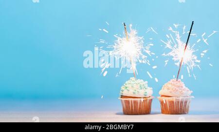 Two cupcakes decorated with colorful sprinkles and sparklers. Festive cupcakes on blue background with copy space left for text or design. Banner Stock Photo