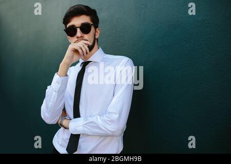 Smartly dressed young businessman standing by a dark wall. Man in formalwear posing in sunglasses. Stock Photo