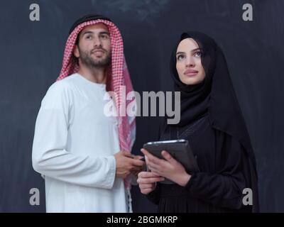 young muslim business couple arabian man with woman in fashionable hijab dress using mobile phone and tablet computer in front of black chalkboard rep Stock Photo