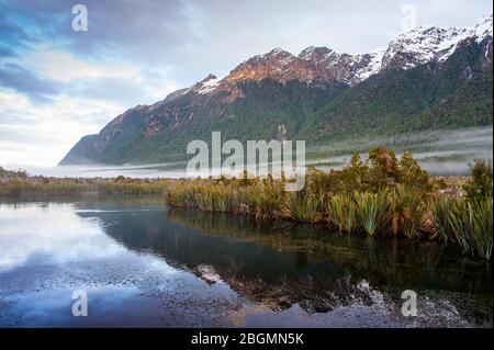 Sunrise, Mirror Lakes in Fiordland National Park, New Zealand. Sunlight falls on snowy mountain tops, reflected in calm, clear water. Stock Photo