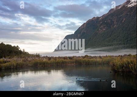 Sunrise, Mirror Lakes in Fiordland National Park, New Zealand. Sunlight falls on snowy mountain tops, reflected in calm, clear water. Stock Photo