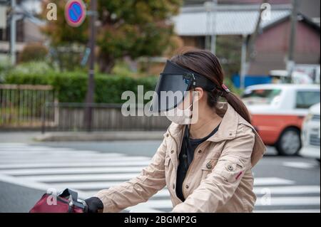 Kyoto, Japan - April 12th, 2019: Young female cyclist on a busy street wearing anti-pollution face mask and a dark visor to shield against the sun. Stock Photo