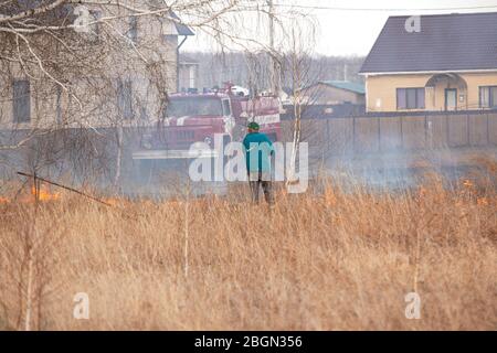 man puts out fire on burning grass. Grass is burning in meadow near private residential building. Azov German national district of Omsk region, Russia Stock Photo