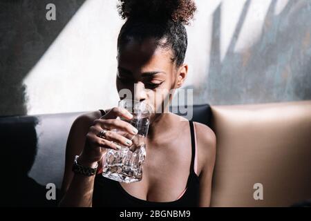 Young black woman with curly hair and tied up drinking water sitting on a black leather sofa Stock Photo
