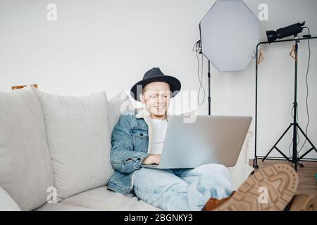 Blogger works laptop, mounts videos for vlog about travel, freelancer photographer in hat video , in background lighting equipment Stock Photo