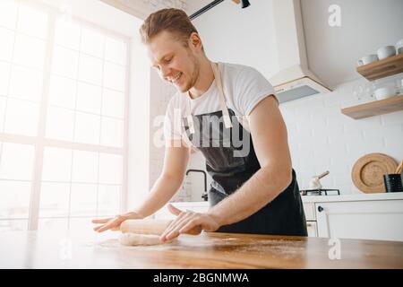 Chef happy man Italian rolls dough on wooden table against of window, light kitchen background Stock Photo