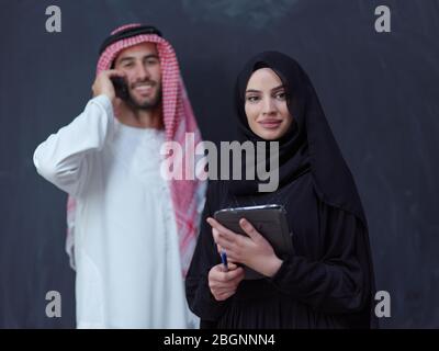young muslim business couple arabian man with woman in fashionable hijab dress using mobile phone and tablet computer in front of black chalkboard rep Stock Photo