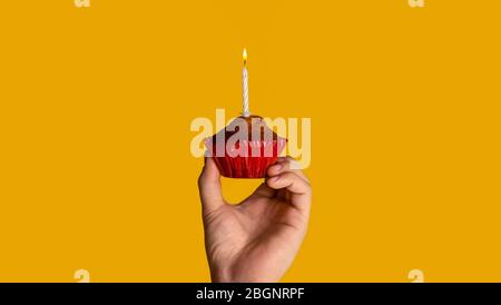 Holiday celebration. Man holding yummy muffin with burning candle on orange background, closeup. Panorama Stock Photo