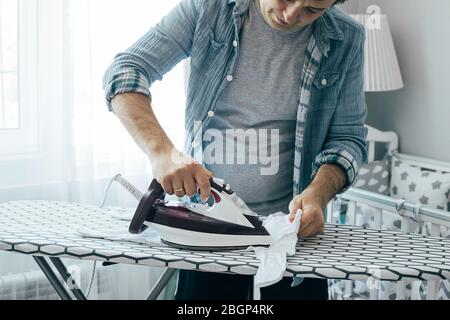 Young father ironing baby clothes in bedroom. Concept of housework, chores at home, domestic life in quarantine period. Stay home. Stock Photo