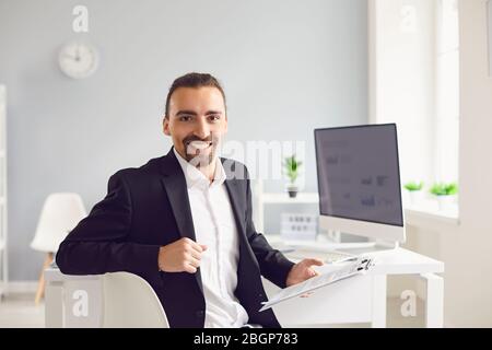 A brunette man in a black jacket sitting at the table works with papers in the office. Stock Photo