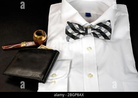 black and white checkered bow tie on folded white dress shirt with wallet and old smoking pipe on black Stock Photo
