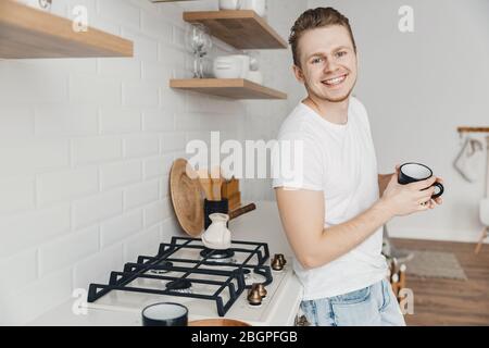 Young man smiles and shares freshly prepared coffee in ceramic black mug. Bright kitchen background Stock Photo
