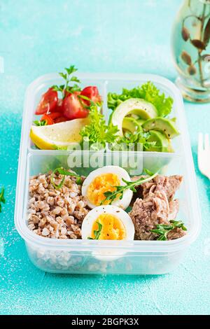 Healthy food. Lunch- Buckwheat porridge, sliced tuna, boiled egg and fresh vegetable salad in container  on blue background. Stock Photo