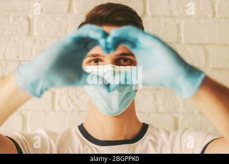 Young man in protective latex gloves and medical face mask gesturing love symbol by hands. Doctor in medical gloves and protective face mask showing h Stock Photo