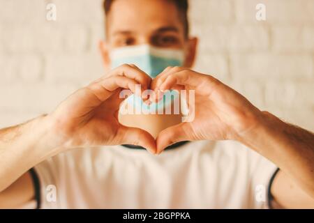 Young man in medical face mask gesturing love shape symbol by hands. Happy doctor in protective mask on face holding fingers in sign of heart. Health Stock Photo