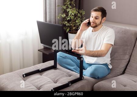 Phone conversation. Stress caucasian gloomy man talking phone working from home office using laptop and internet. Remote work in stress, freelancer Stock Photo