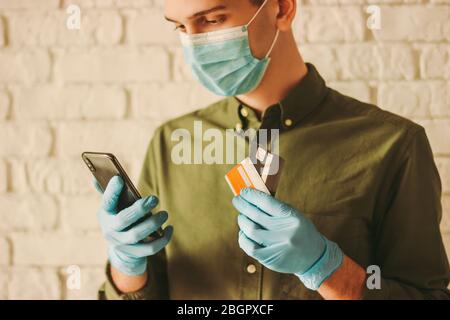 Hipster man in protective face mask and medical gloves shopping in online store. Young man in medical face mask, protective gloves holding mobile phon Stock Photo