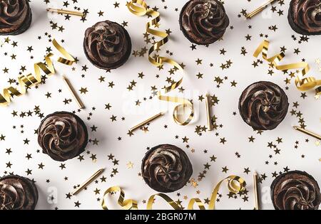 Chocolate cupcakes with chocolate icing and sprinkled gold sparkles on white background decorated with golden star shaped confetti, candles and party Stock Photo