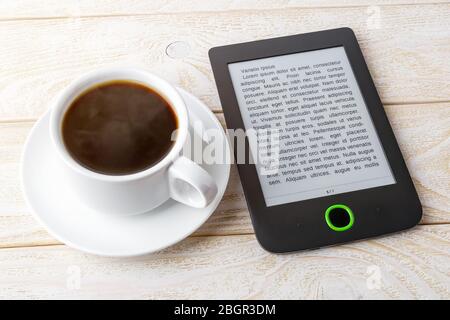 Copy space on a screen of e-book reader near cup of hot coffee on a white wooden table. E-reading for pleasure and education. Front view. Stock Photo