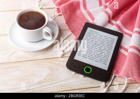 Cup of hot coffee near e-book reader over red cotton stole on a white wooden surface. E-reading for pleasure and education. Top view. Stock Photo
