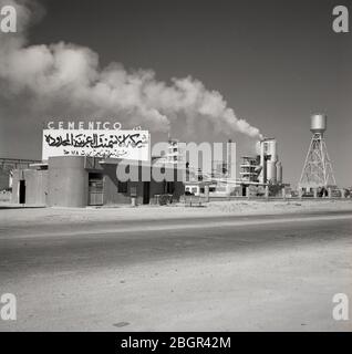 1960s, Saudi Arabia, exterior view of Cementco, a Saudi cement factory, in Al Hofuf, showing smoke coming out of a chimney and a sign in Arabic and in English above at the entrance. Stock Photo