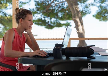 Young female photographer checking and editing photos on her laptop computer, sitting at a desk outside in summer resort. Stock Photo