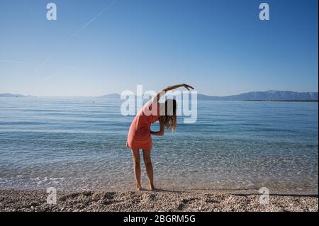 View from behind of a young woman standing ankle deep in calm morning sea water stretching with her arm to the side. Stock Photo