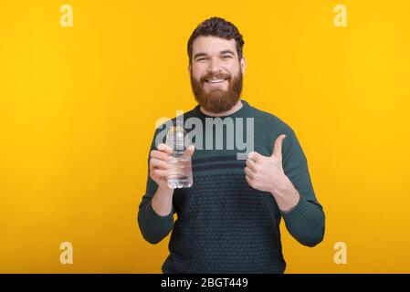 Photo of young man standing over yellow background and holding water bottle and showing thumbs up Stock Photo