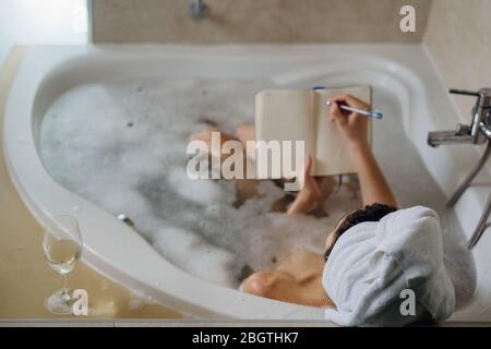 Calm woman enjoying a relaxing bath at home.Spa self care night.Inspired creative person writing gratitude diary/journal.Resolution list.Making plans. Stock Photo