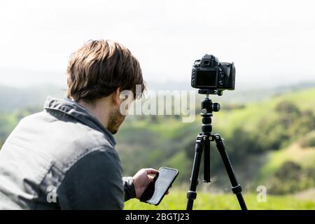 Person using smartphone app to control camera on tripod outdoors Stock Photo