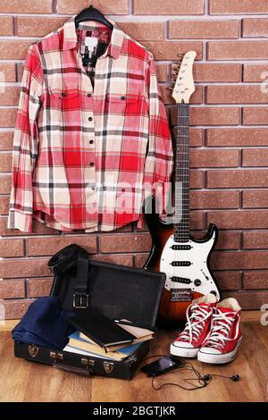 Guitar, clothes, boots and suitcase with books on floor on brick wall background Stock Photo