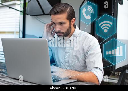 Hologram projections of symbols from a computer shoot around a man with brown slicked hair working on laptop on a picnic table remotely near his campe Stock Photo