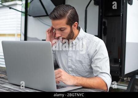 A man wearing a long sleeve grey button down business shirt with brown slicked comb over hair working on laptop on a picnic table remotely near his ca Stock Photo