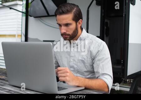 A man wearing a long sleeve grey button down business shirt with brown slicked comb over hair working on laptop on a picnic table remotely near his ca Stock Photo