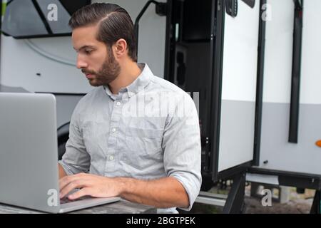 A man wearing a long sleeve grey button down business shirt with brown slicked comb over hair working on laptop on a picnic table remotely near his ca Stock Photo